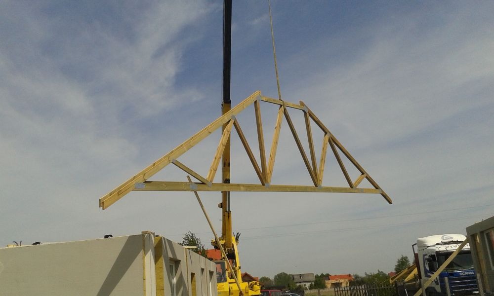 Montaż drewnianej więźby dachowej za pomocą dźwigu na budowie domu modułowego, widoczny fragment ściany z oknem.