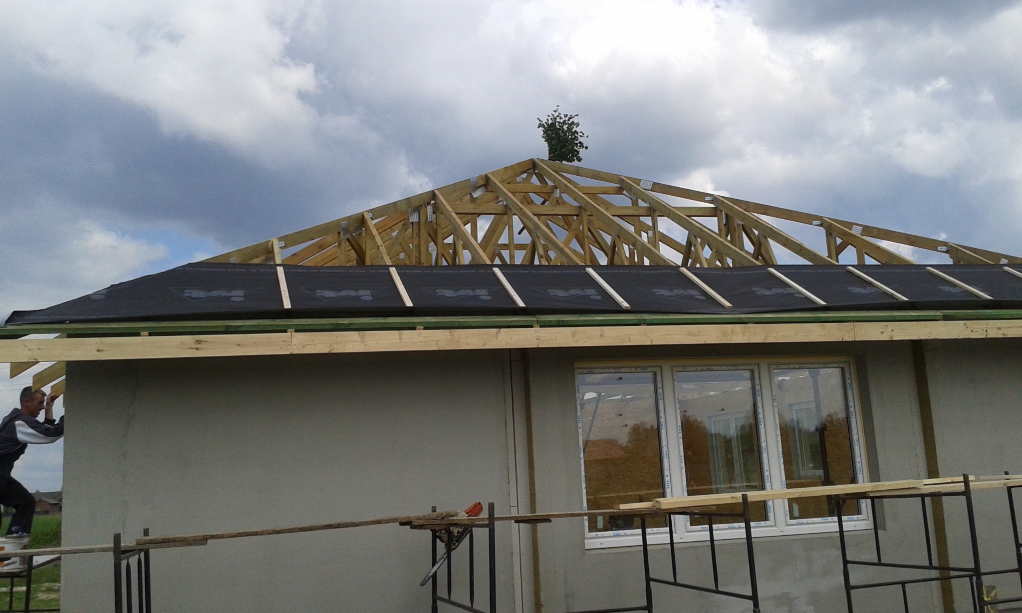 Konstrukcja dachu domu murowanego w trakcie budowy, widoczny pracownik na rusztowaniu, membrana dachowa i drewniane elementy więźby, małe drzewko na szczycie dachu.