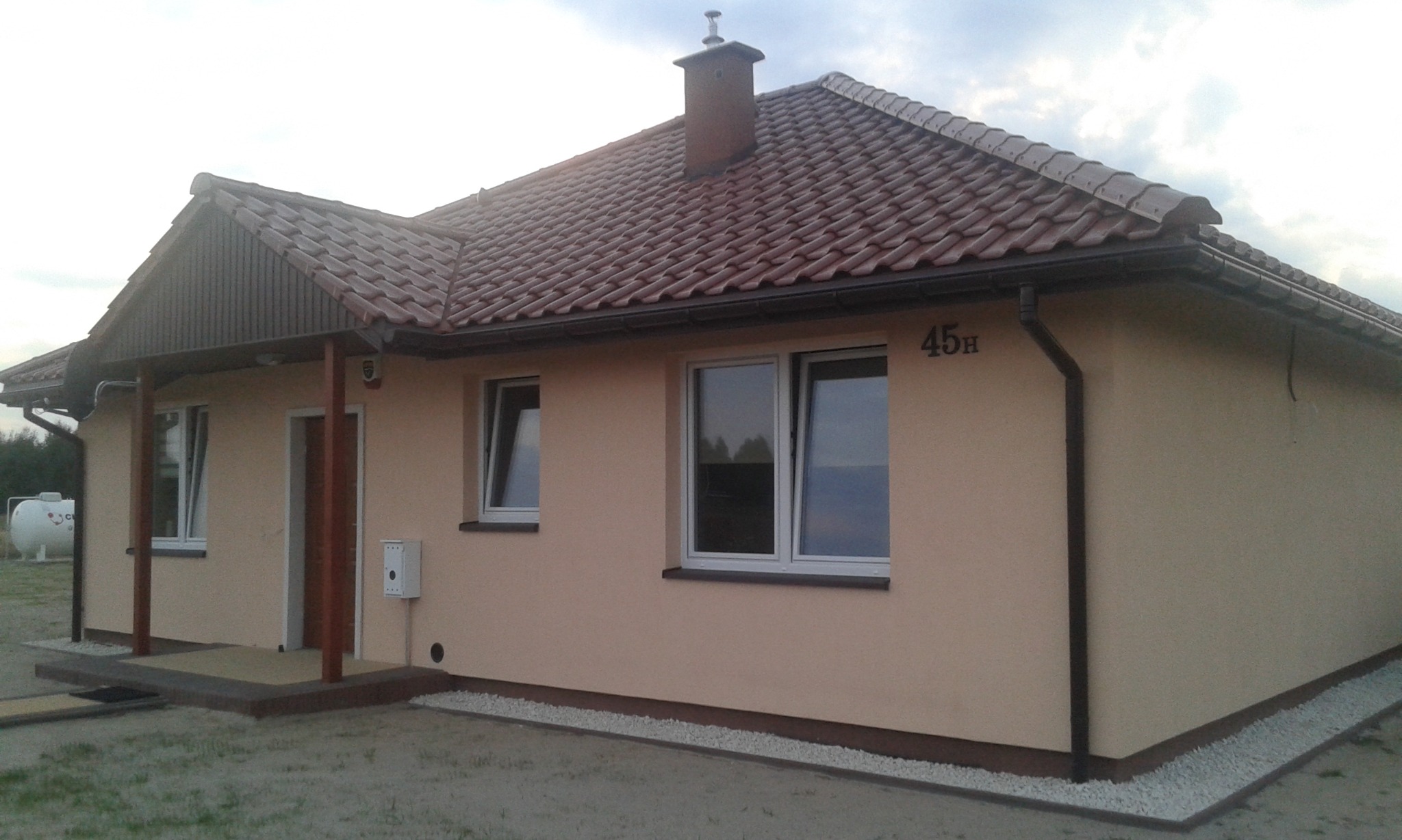 Parterowy dom z beżową elewacją, brązową dachówką i numerem 45H nad oknem, widoczny zbiornik na gaz w tle.