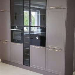 NOWY WYMIAR MEBLI AGATA PIECHOTA - Nowoczesna zabudowa kuchenna w odcieniach szarości z czarnymi szklanymi frontami, wbudowanymi piekarnikami i minimalistycznymi złotymi uchwytami, oświetlenie sufitowe punktowe.