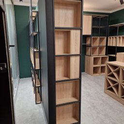 NOWY WYMIAR MEBLI AGATA PIECHOTA - Nowoczesne regały modułowe z dębowymi półkami i czarnymi metalowymi ramami, ustawione w showroomie z zielonymi ścianami i szarą podłogą.