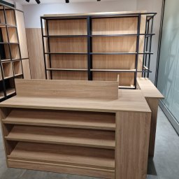 NOWY WYMIAR MEBLI AGATA PIECHOTA - Jasna, drewnopodobna zabudowa sklepowa z otwartymi półkami i czarnymi, metalowymi regałami w tle, oświetlona punktowymi lampami sufitowymi.