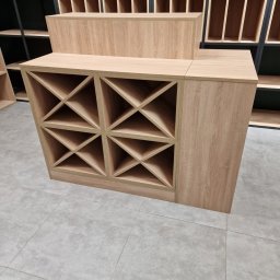 NOWY WYMIAR MEBLI AGATA PIECHOTA - Drewniana, modułowa konstrukcja z półkami na wino, w tle regał z otwartymi przegródkami, całość w jasnej kolorystyce.