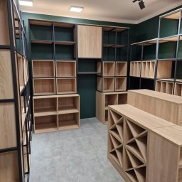 NOWY WYMIAR MEBLI AGATA PIECHOTA - Wnętrze sklepu z otwartymi regałami i półkami z jasnego drewna, z czarnymi metalowymi ramami na tle ciemnozielonych ścian, oświetlone punktowymi lampami sufitowymi.