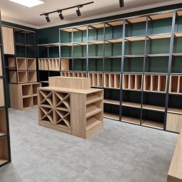 NOWY WYMIAR MEBLI AGATA PIECHOTA - Wnętrze sklepu z pustymi półkami z jasnego drewna i czarnymi metalowymi ramami na tle ciemnozielonej ściany, oświetlone punktowymi reflektorami.