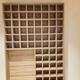 NOWY WYMIAR MEBLI AGATA PIECHOTA - Drewniana konstrukcja z licznymi kwadratowymi przegródkami, częściowo zamknięta panelem z trzema poziomymi, złotymi prętami, tworząca regał o asymetrycznym układzie.