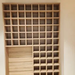 Drewniana konstrukcja z licznymi kwadratowymi przegr&oacute;dkami, częściowo zamknięta panelem z trzema poziomymi, złotymi prętami, tworząca regał o asymetrycznym układzie.