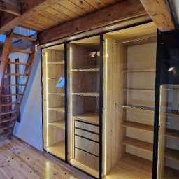 NOWY WYMIAR MEBLI AGATA PIECHOTA - Nowoczesna szafa wnękowa z przesuwnymi, szklanymi drzwiami, oświetleniem LED i drewnianymi półkami, umieszczona na poddaszu ze schodami i drewnianą podłogą.