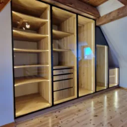 Szafa wnękowa na poddaszu z drewnianymi elementami, czarnymi ramami i oświetleniem LED, dopasowana do skosu dachu, z widoczną podłogą z jasnego drewna.