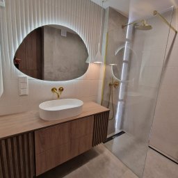 NOWY WYMIAR MEBLI AGATA PIECHOTA - Nowoczesna łazienka z drewnianą szafką pod umywalką, owalnym lustrem z podświetleniem LED oraz kabiną prysznicową ze złotymi elementami.