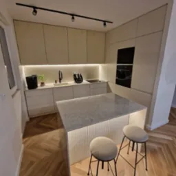 Nowoczesna kuchnia z wyspą oświetloną od dołu, jasne fronty szafek z delikatnym frezowaniem, drewniana podłoga ułożona w jodełkę, czarne oświetlenie szynowe na suficie.