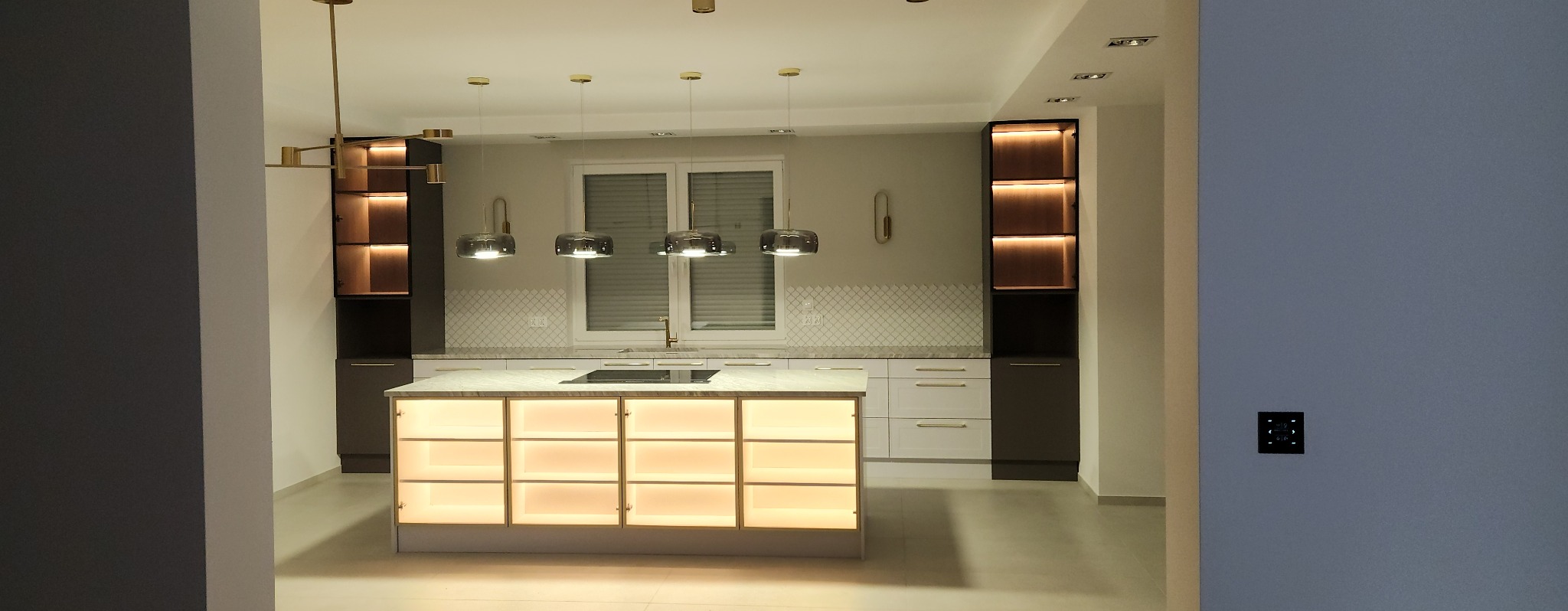 Nowoczesna kuchnia z wyspą oświetloną od wewnątrz, wiszące lampy z szarymi kloszami, zabudowa w kolorach bieli i grafitu z oświetlonymi półkami.