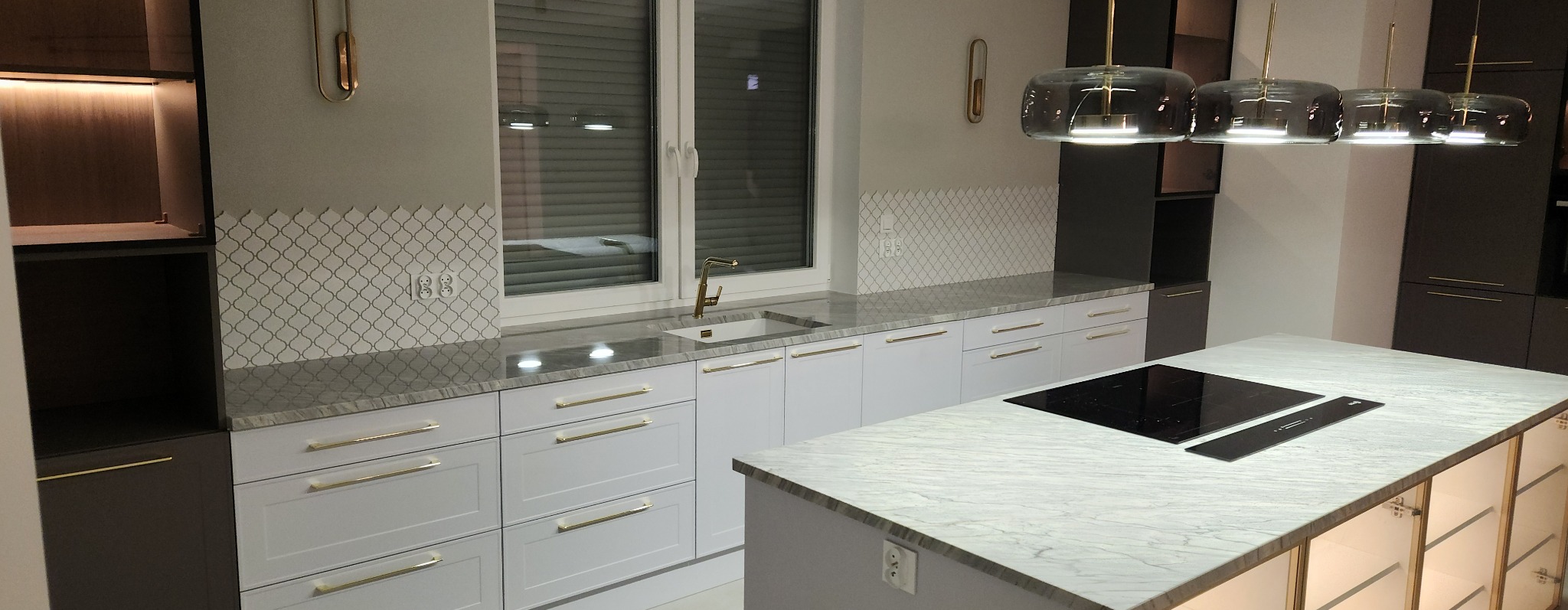 Nowoczesna kuchnia z białymi szafkami, złotymi uchwytami, marmurowym blatem i wyspą kuchenną z płytą indukcyjną, oświetlona wiszącymi lampami w stylu industrialnym.
