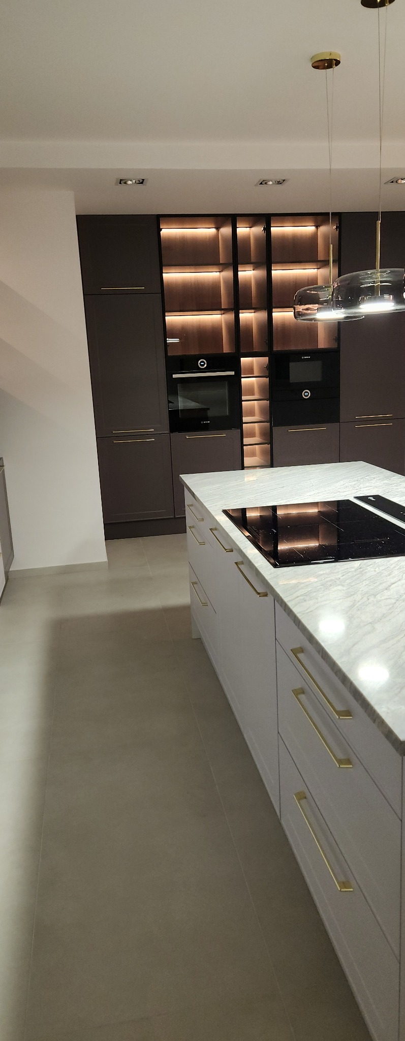 Nowoczesna kuchnia z wyspą, fronty w kolorze białym z złotymi uchwytami, zabudowa w kolorze brązowym z oświetleniem LED, dwie czarne wbudowane kuchenki.