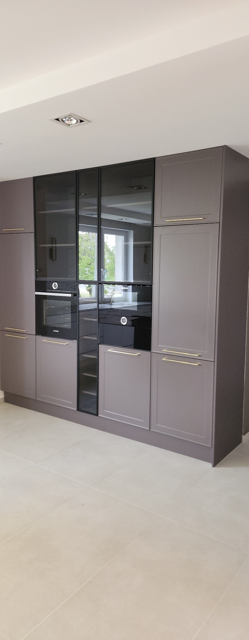 Nowoczesna zabudowa kuchenna w odcieniach szarości z czarnymi szklanymi frontami, wbudowanymi piekarnikami i minimalistycznymi złotymi uchwytami, oświetlenie sufitowe punktowe.