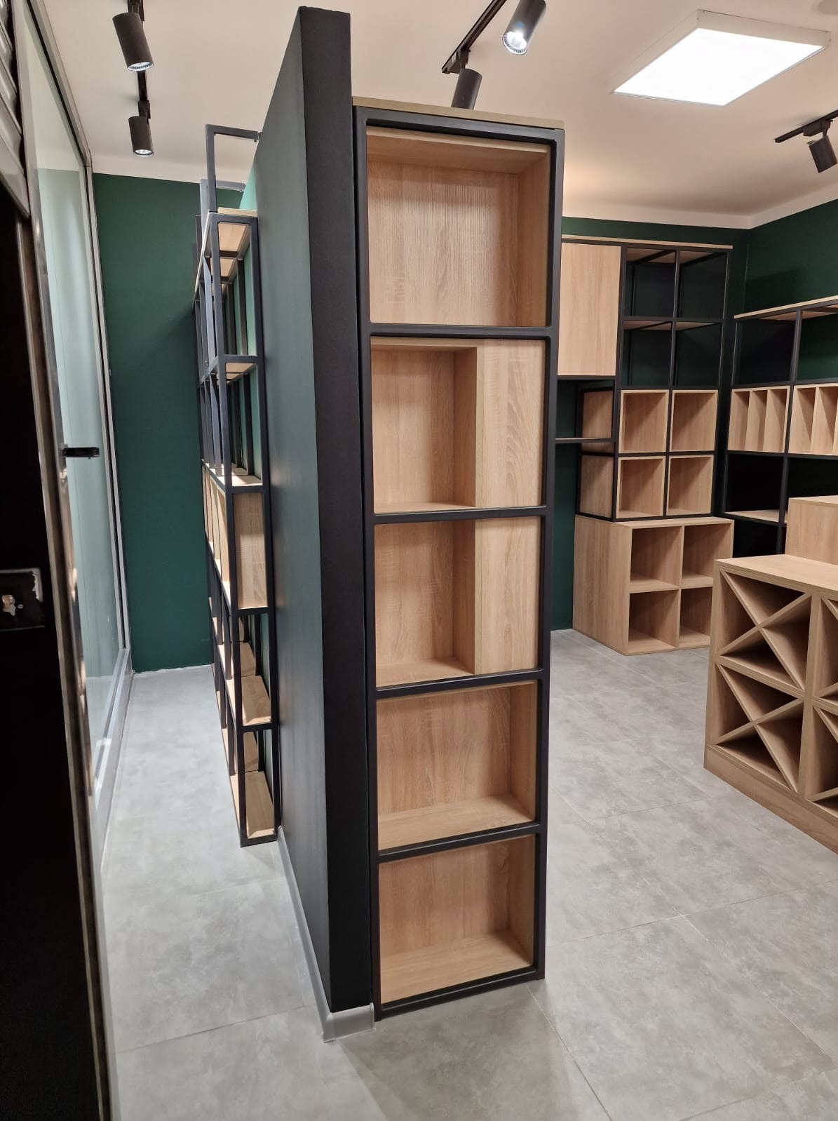 Nowoczesne regały modułowe z dębowymi półkami i czarnymi metalowymi ramami, ustawione w showroomie z zielonymi ścianami i szarą podłogą.