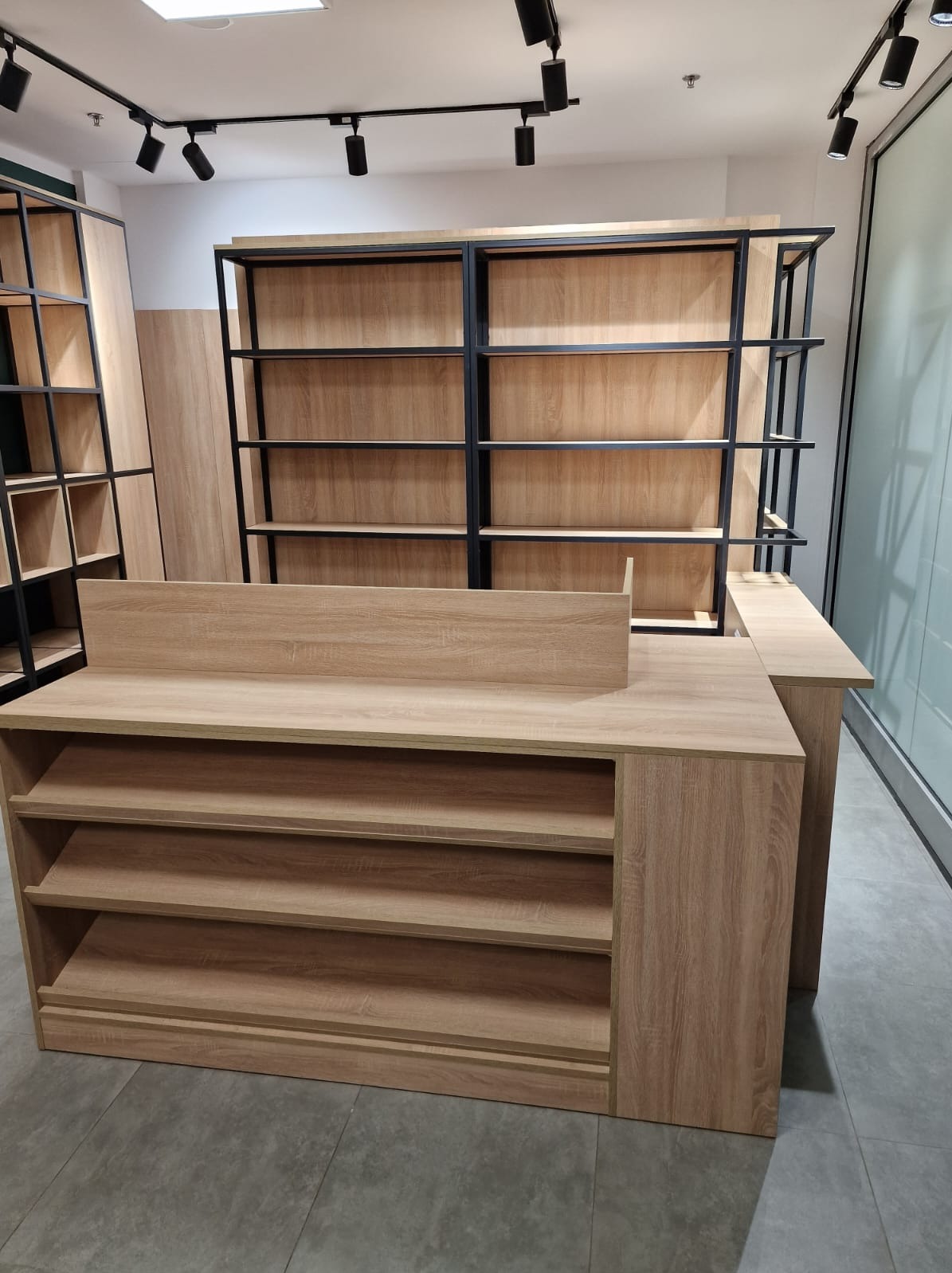 Jasna, drewnopodobna zabudowa sklepowa z otwartymi półkami i czarnymi, metalowymi regałami w tle, oświetlona punktowymi lampami sufitowymi.