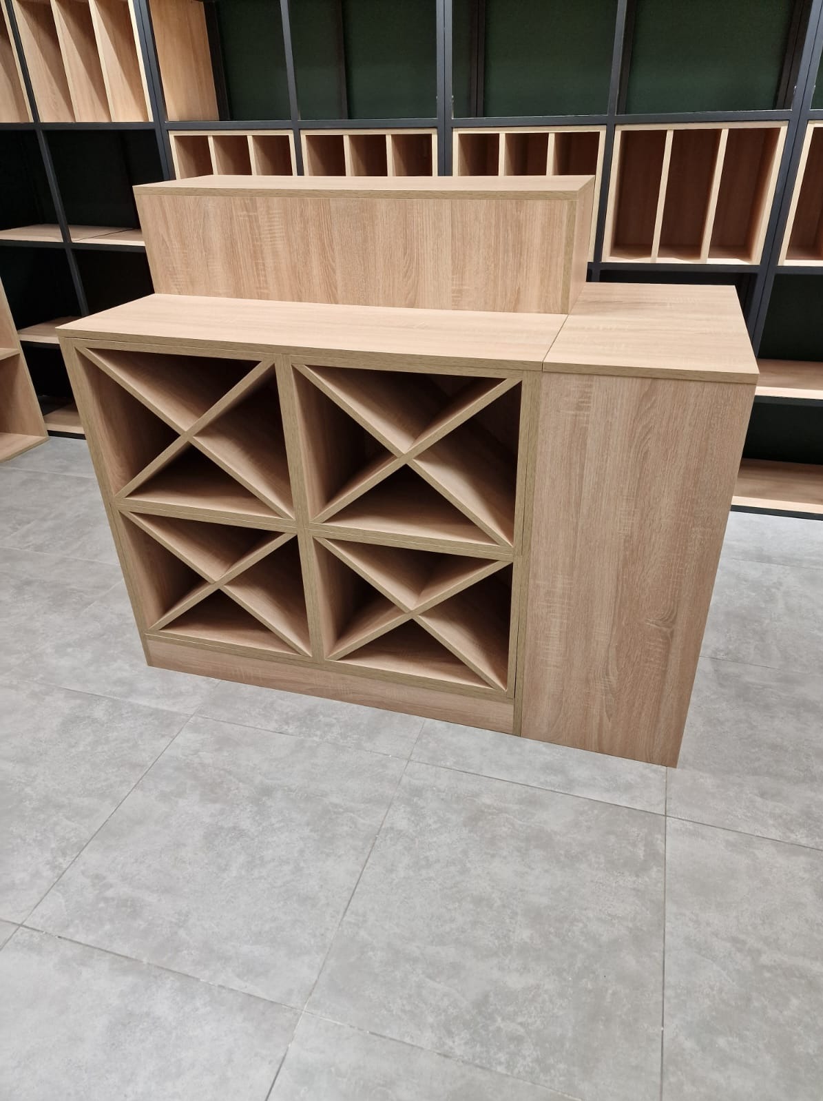 Drewniana, modułowa konstrukcja z półkami na wino, w tle regał z otwartymi przegródkami, całość w jasnej kolorystyce.