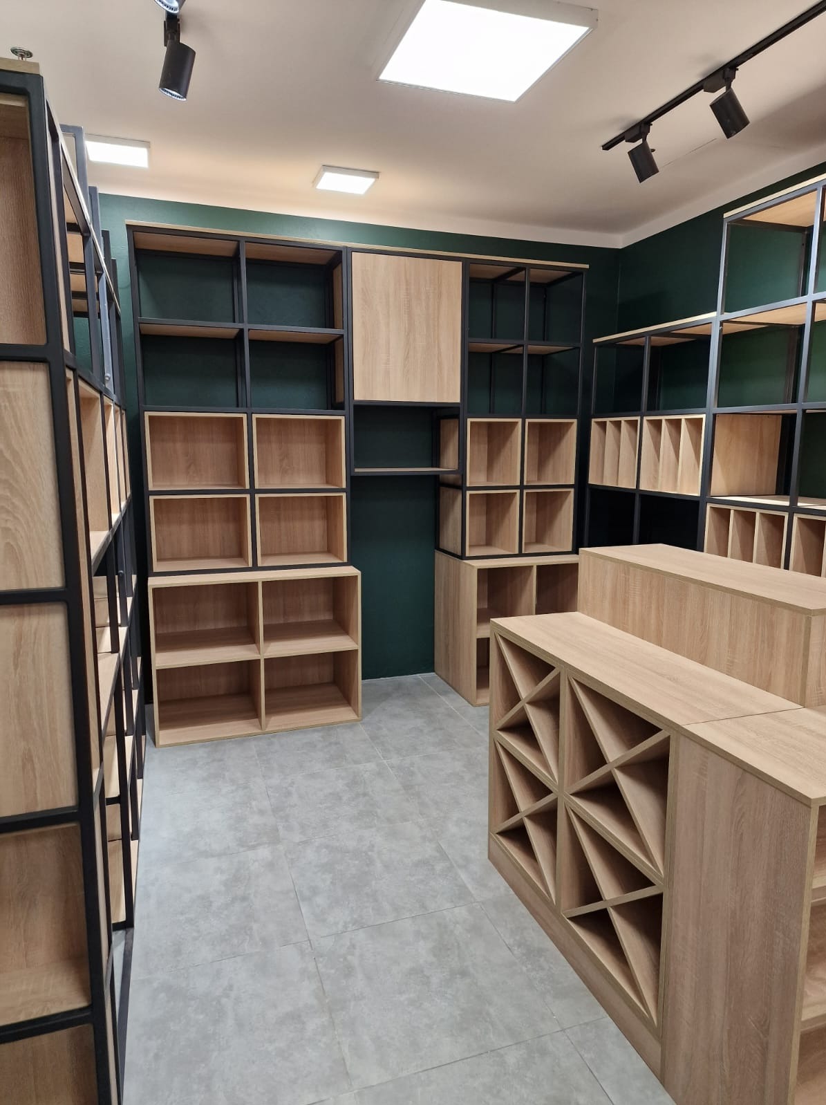 Wnętrze sklepu z otwartymi regałami i półkami z jasnego drewna, z czarnymi metalowymi ramami na tle ciemnozielonych ścian, oświetlone punktowymi lampami sufitowymi.