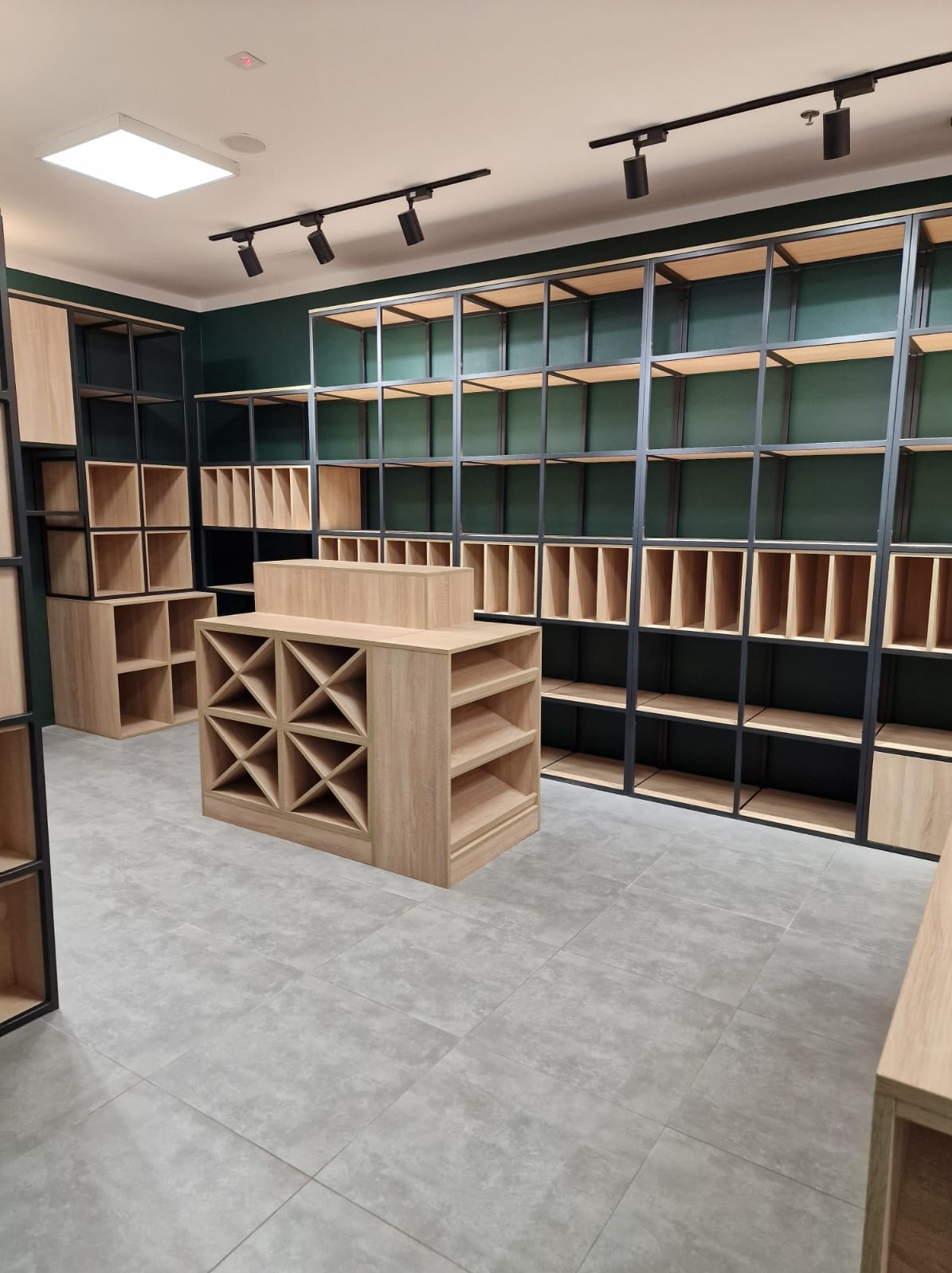 Wnętrze sklepu z pustymi półkami z jasnego drewna i czarnymi metalowymi ramami na tle ciemnozielonej ściany, oświetlone punktowymi reflektorami.