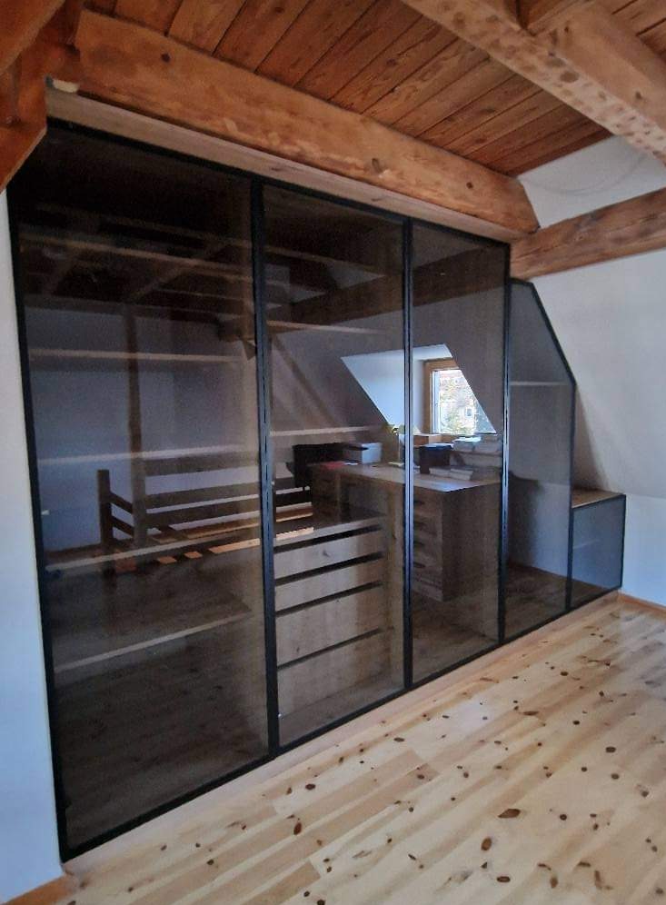 Szafa wnękowa z przesuwnymi, szklanymi drzwiami w czarnej ramie, dopasowana do skosu poddasza, z drewnianymi półkami i szufladami, widoczna na tle drewnianej podłogi i belek stropowych.