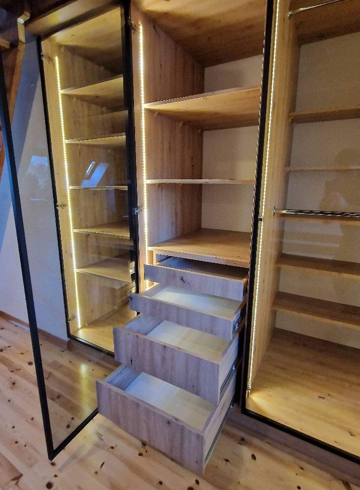 Wnętrze drewnianej szafy z oświetleniem LED, z trzema wysuniętymi szufladami i półkami, widoczne okucia szuflad.