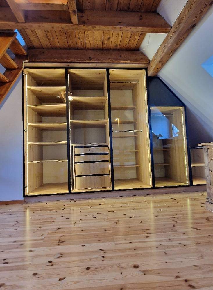 Drewniana zabudowa wnęki na poddaszu z szafami z oświetleniem LED, widoczne półki i szuflady, podłoga z jasnego drewna.