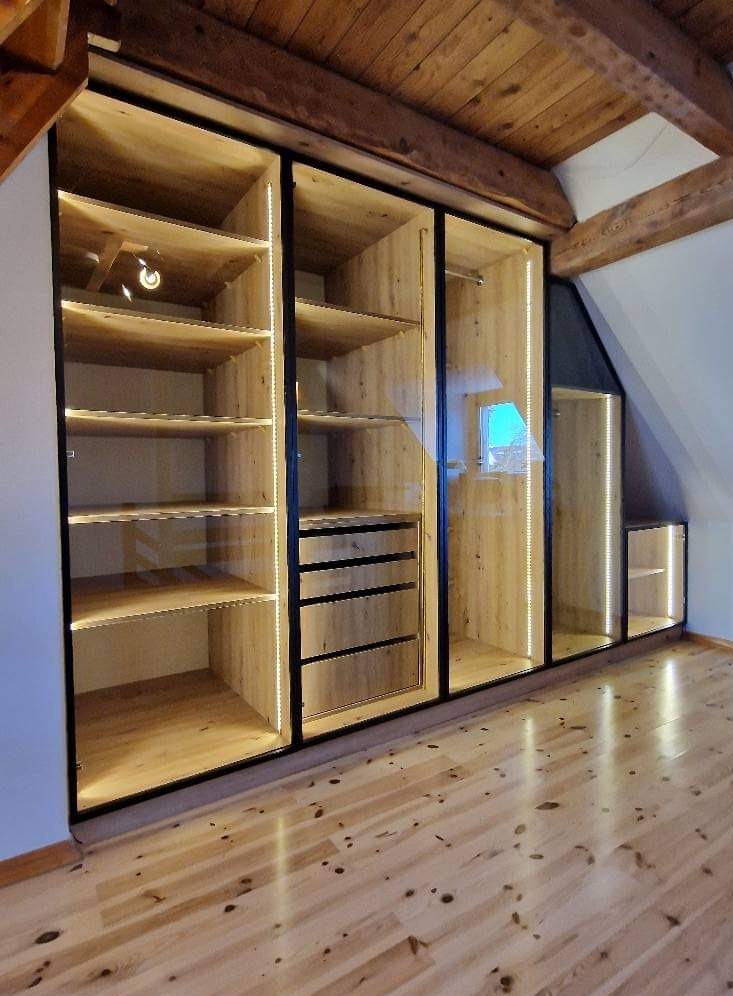 Wnętrze poddasza z drewnianą podłogą i zabudowaną szafą z przesuwnymi, szklanymi drzwiami, podświetloną ledami, z półkami i szufladami.