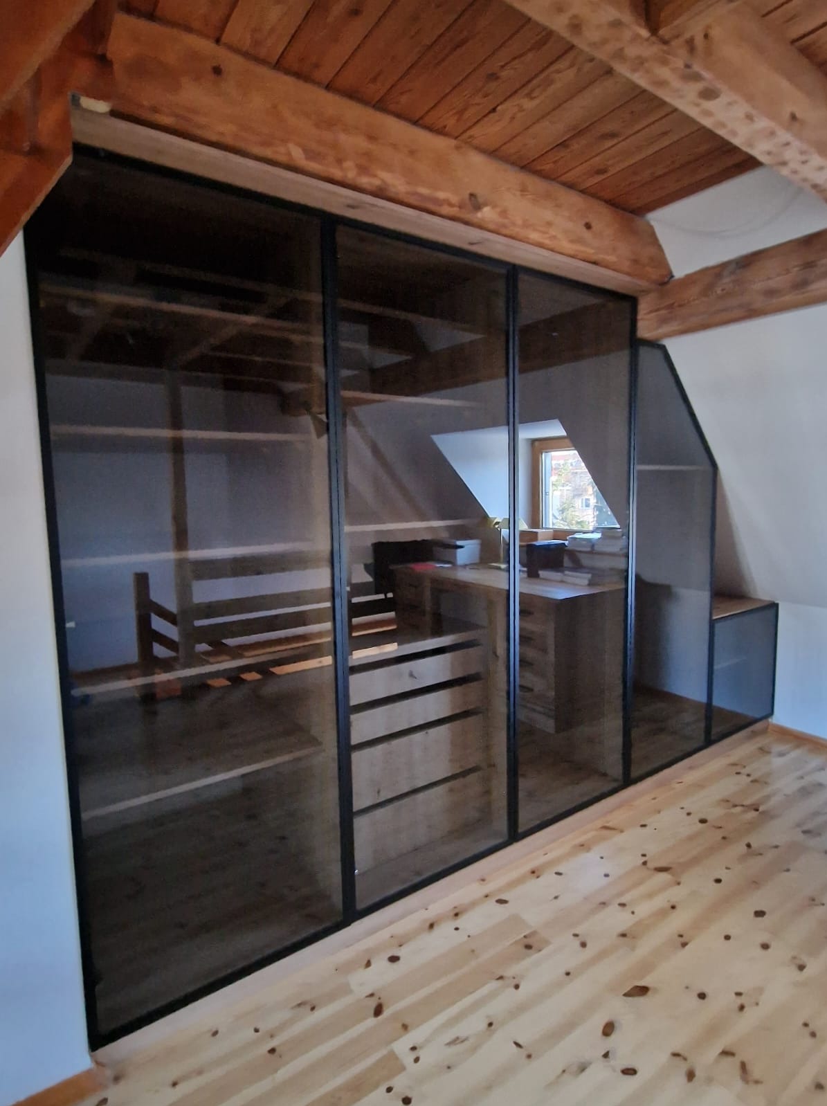 Przeszklona szafa wnękowa z czarnymi ramami, wkomponowana w skośny sufit poddasza, z widocznym drewnianym regałem i biurkiem wewnątrz.