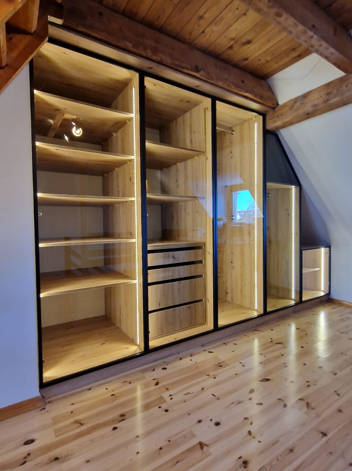 Szafa wnękowa na poddaszu z drewnianymi elementami, czarnymi ramami i oświetleniem LED, dopasowana do skosu dachu, z widoczną podłogą z jasnego drewna.