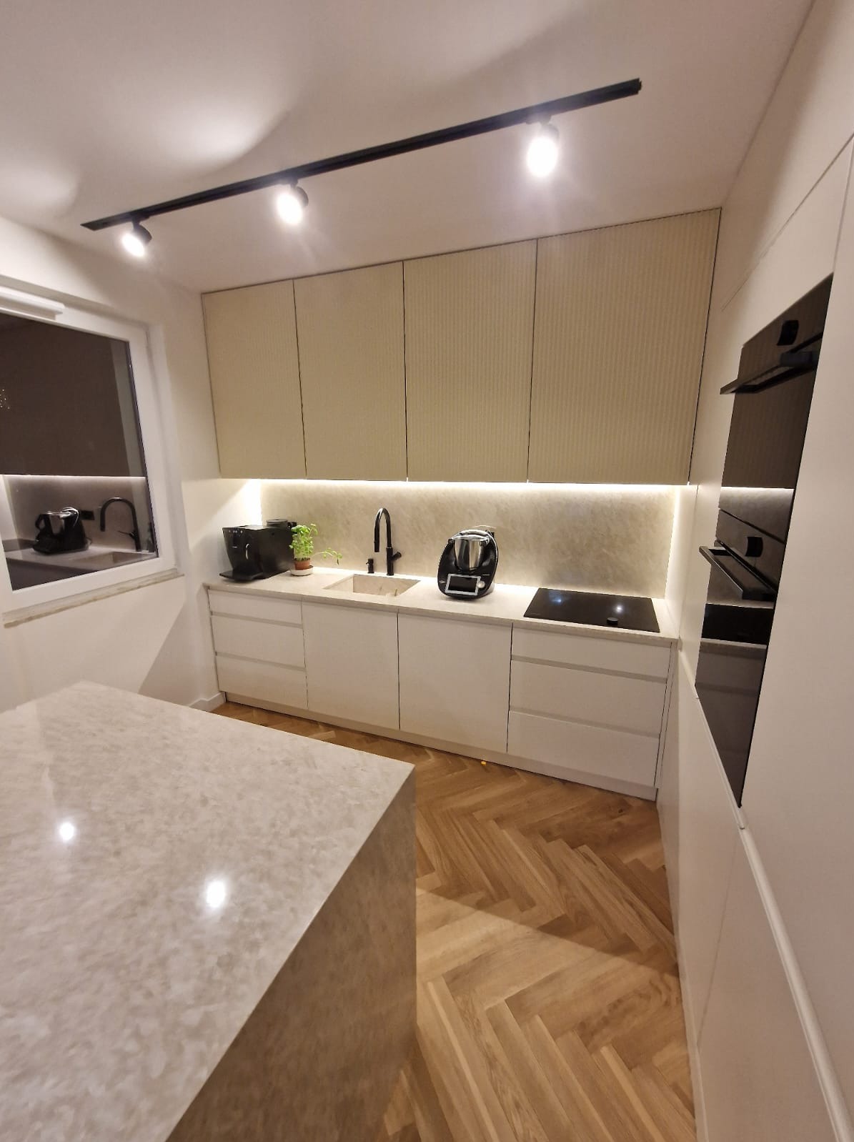 Nowoczesna kuchnia z białymi szafkami, ryflowanymi frontami górnych szafek, blatem z kamienia i drewnianą podłogą w jodełkę.