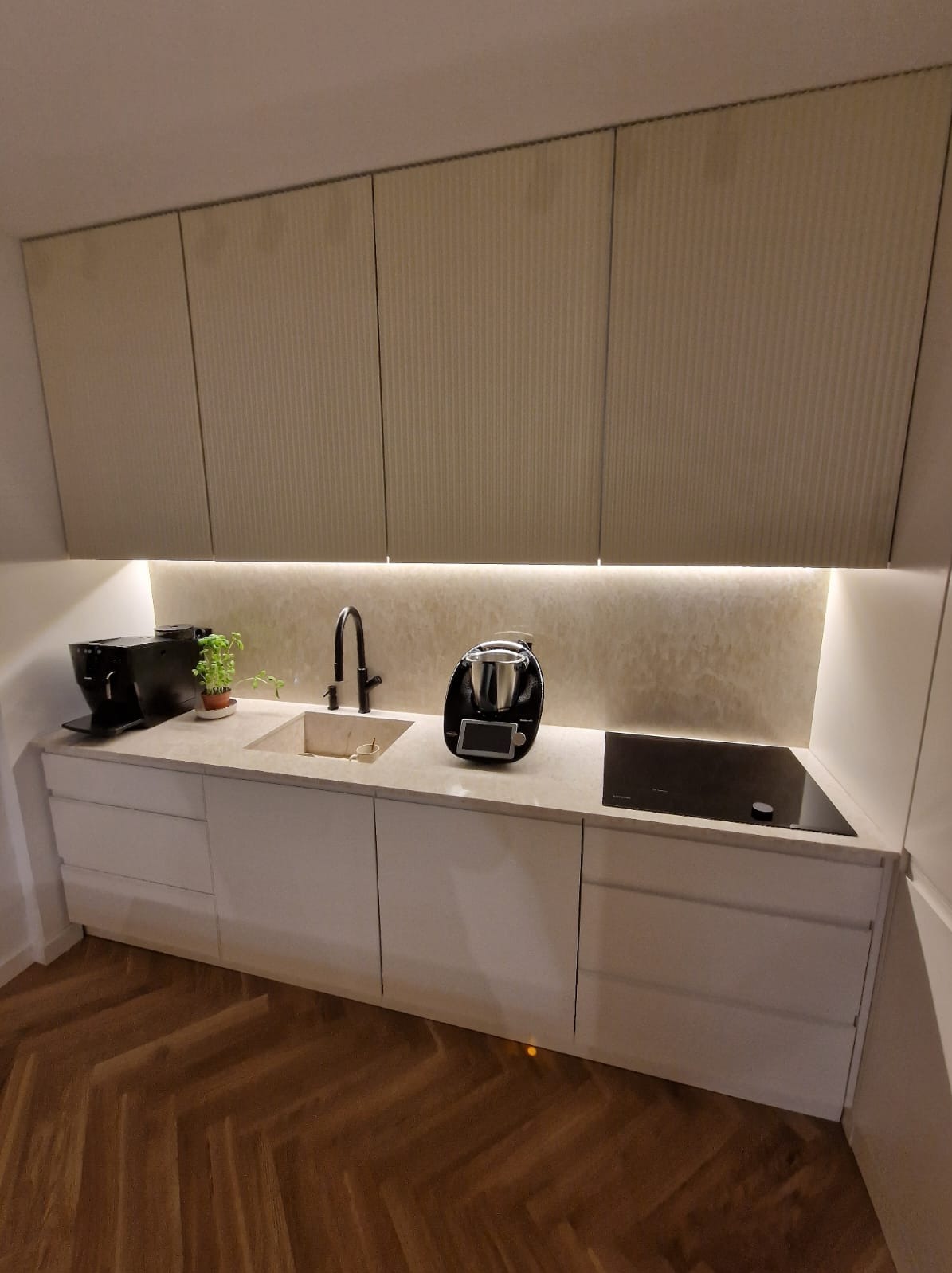 Nowoczesna kuchnia z jasnymi, ryflowanymi szafkami wiszącymi, kamiennym blatem, czarną armaturą, płytą indukcyjną i sprzętami AGD, oświetlona listwą LED.