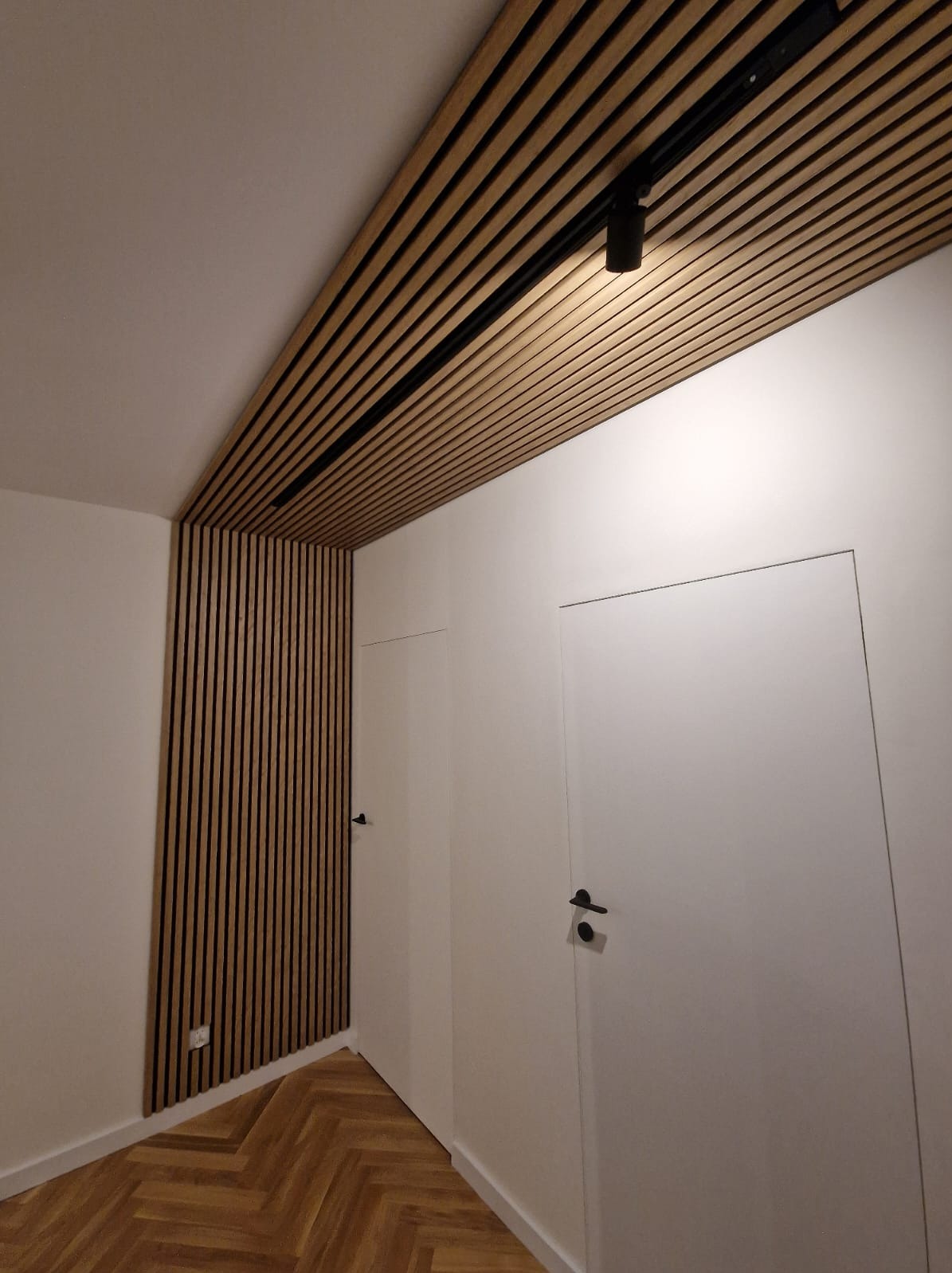 Nowoczesne wnętrze z drewnianymi lamelami na suficie i ścianie, oświetleniem punktowym na szynie, parkietem w jodełkę i minimalistycznymi białymi drzwiami.