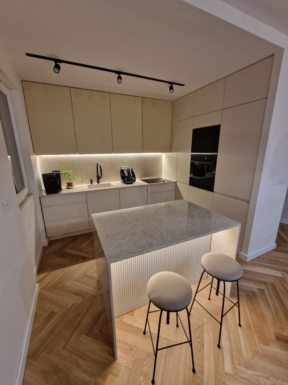 Nowoczesna kuchnia z wyspą oświetloną od dołu, jasne fronty szafek z delikatnym frezowaniem, drewniana podłoga ułożona w jodełkę, czarne oświetlenie szynowe na suficie.