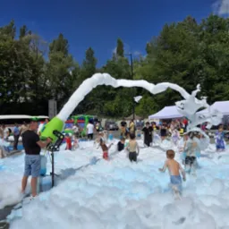 Dzieci bawiące się w pianie wytwarzanej przez zielony generator piany na tle drzew i namiotów.