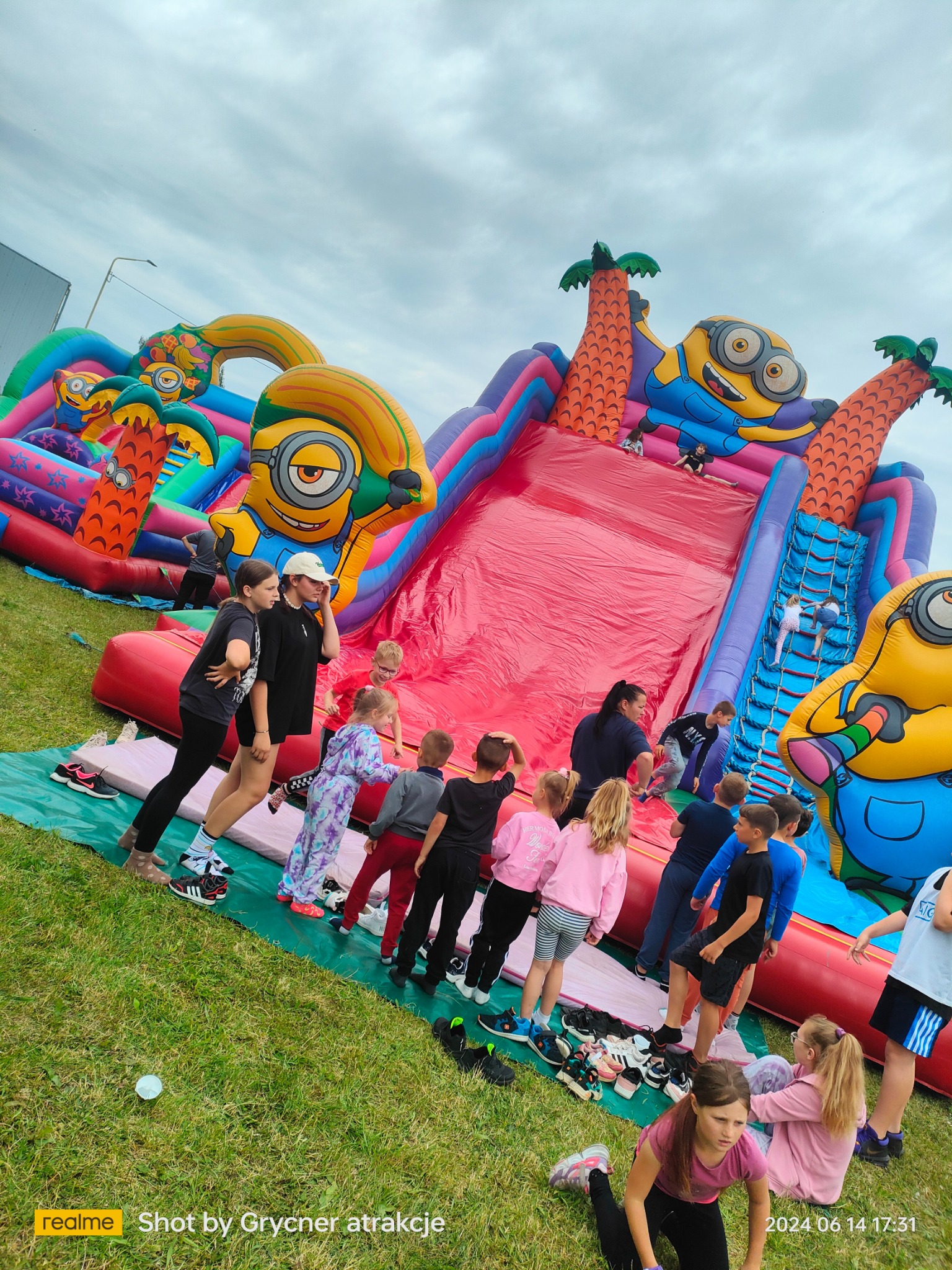 Kolorowa dmuchana zjeżdżalnia z motywem Minionków, otoczona grupą dzieci czekających na swoją kolej, buty zdjęte i ułożone obok, pochmurne niebo w tle.