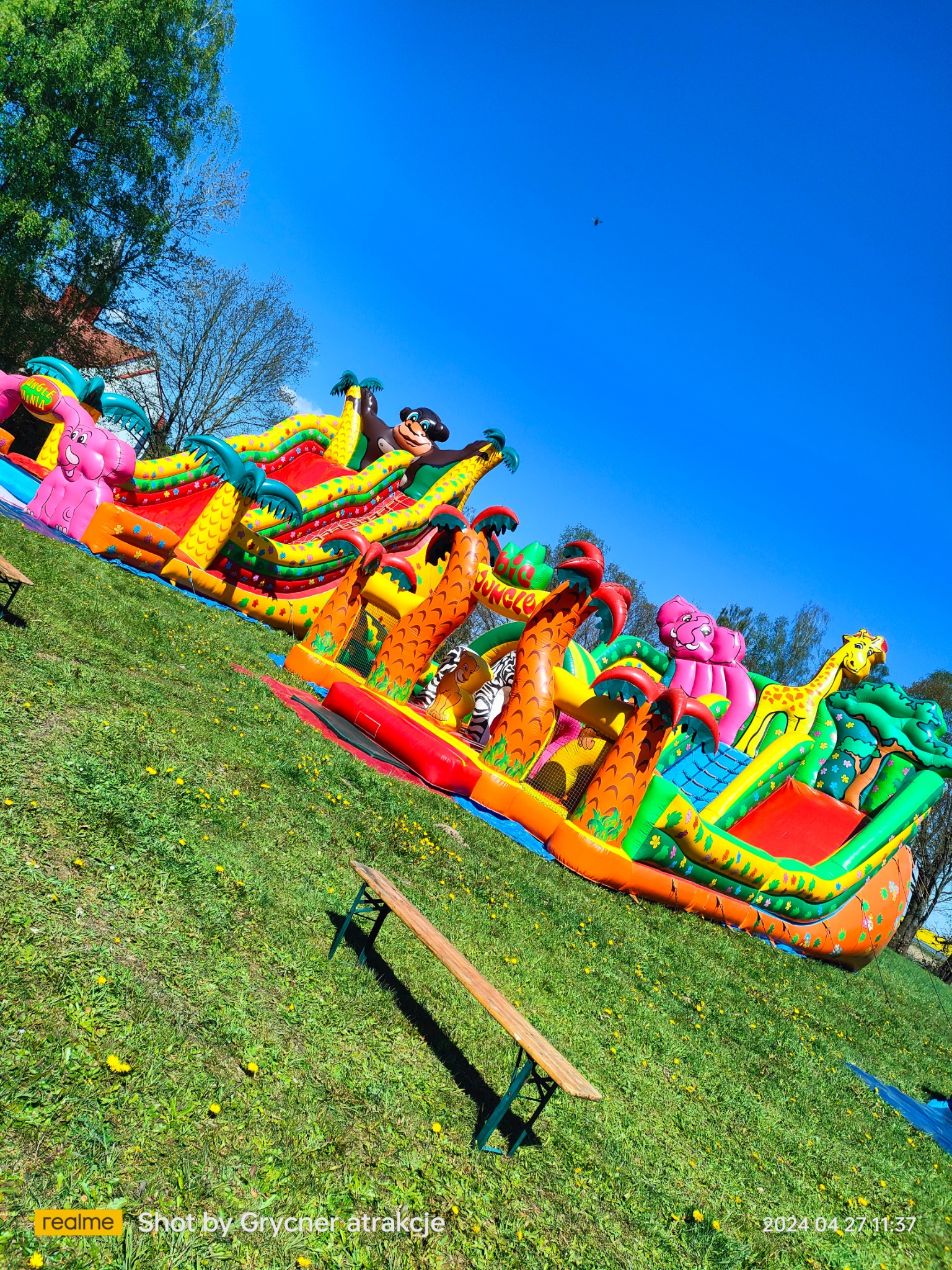 Kolorowy, dmuchany plac zabaw w motywie dżungli z palmami, słoniem, małpą i żyrafą na trawiastym terenie z żółtymi kwiatami pod błękitnym niebem, widziany z boku, z fragmentem drewnianej ławki...