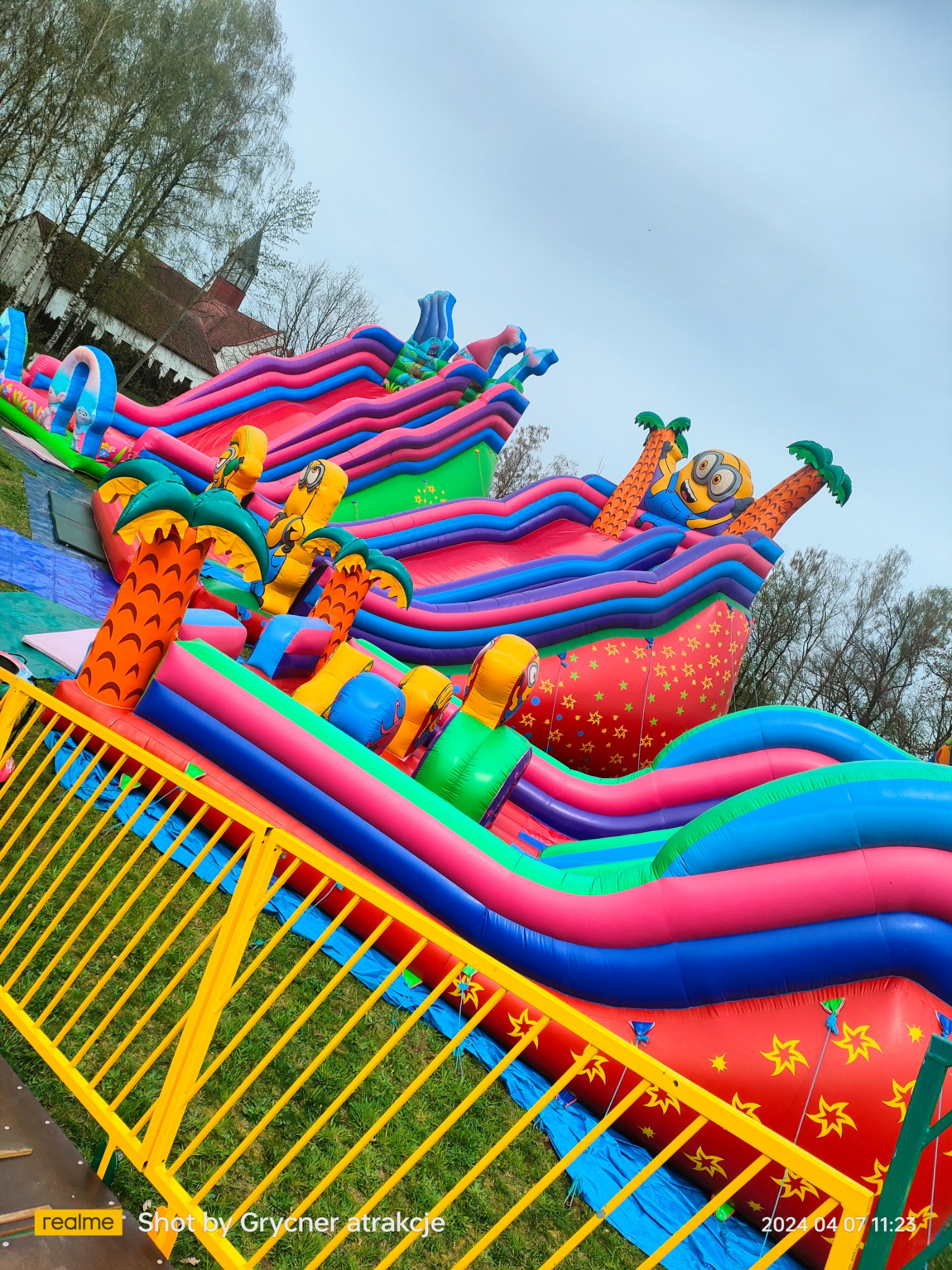 Kolorowy dmuchany zamek ze zjeżdżalniami, motywem palm i postacią przypominającą Minionka, zabezpieczony żółtą barierką, na tle trawnika i budynku z czerwonym dachem.