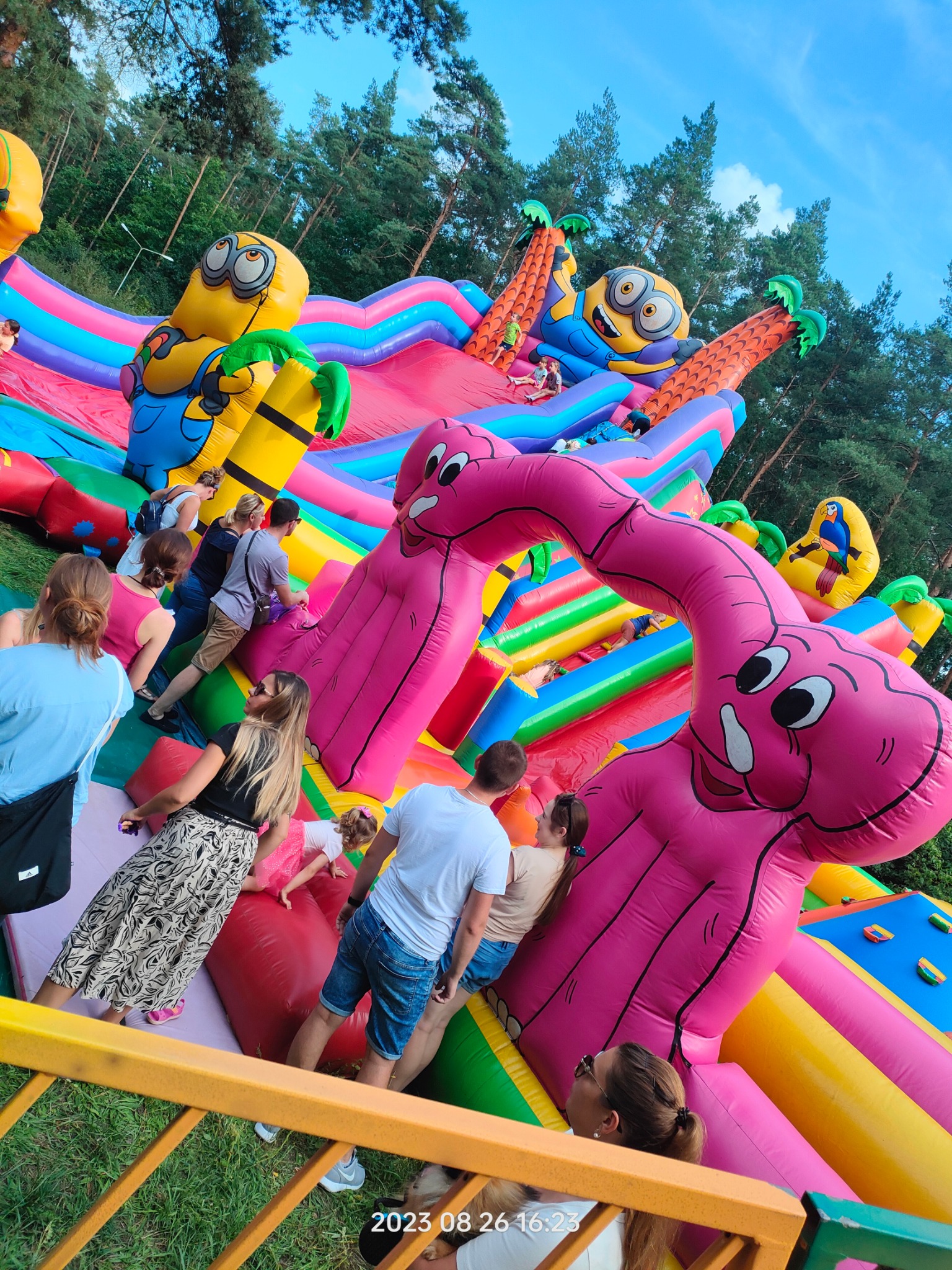 Kolorowy, dmuchany plac zabaw z motywami Minionków i różowych flamingów, widziany z dołu, z bawiącymi się dziećmi i dorosłymi w tle drzew