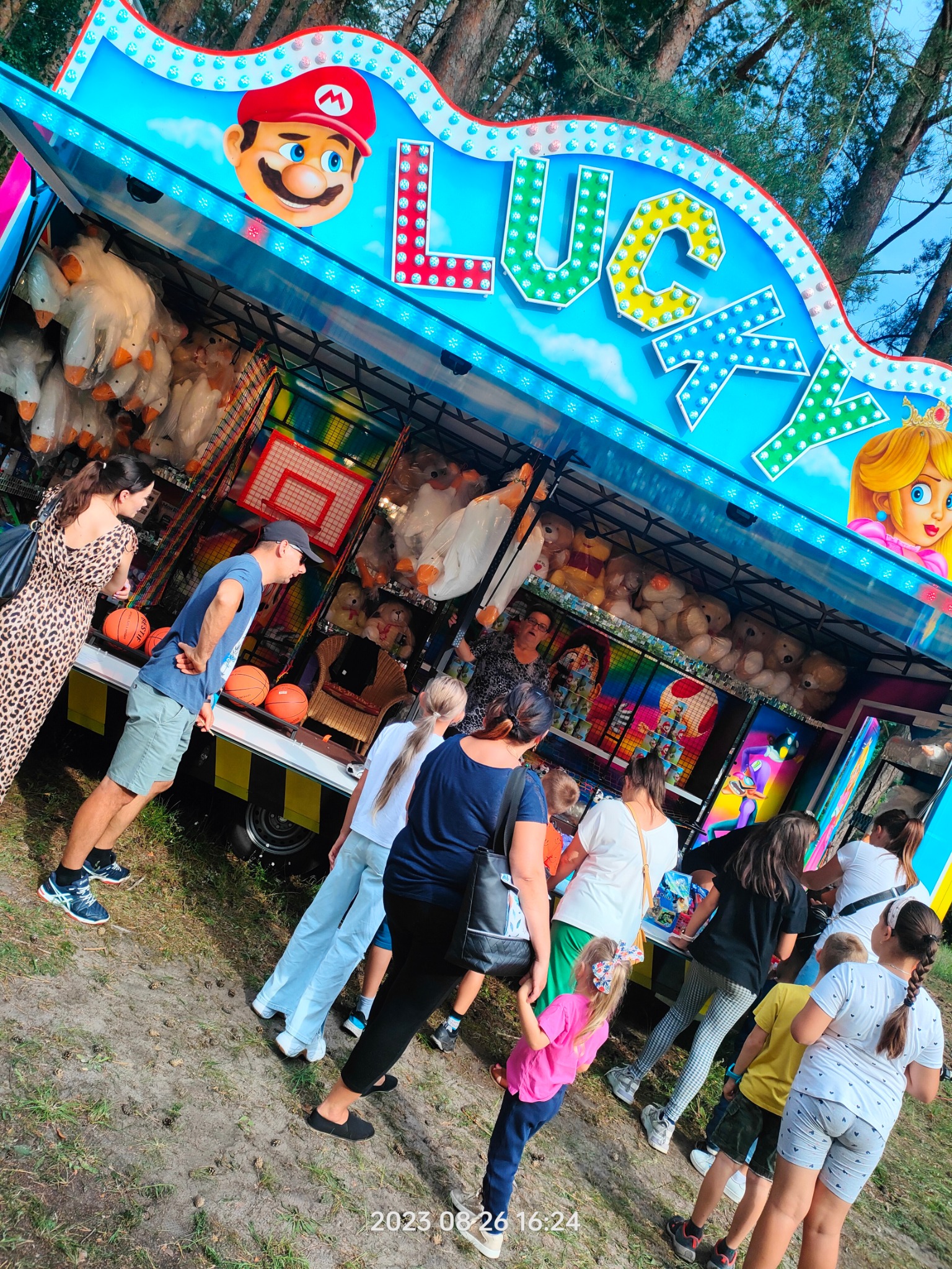 Kolorowy wóz z grami zręcznościowymi i pluszakami, z wizerunkami postaci z gier, otoczony grupą osób, w tym dzieci, próbujących swoich sił w grach.