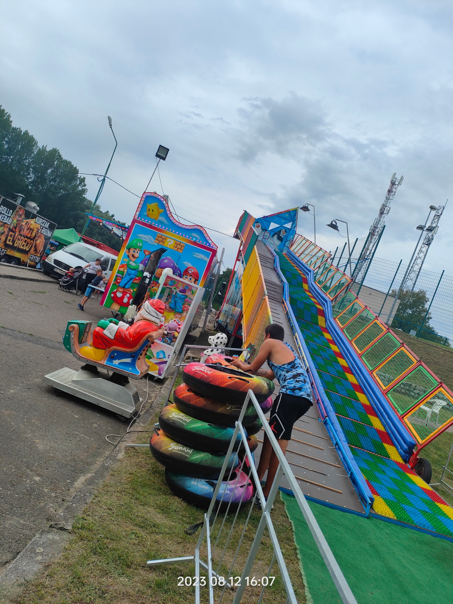 Kolorowa dmuchana zjeżdżalnia z motywem Mario i platforma z karuzelą w kształcie sań Świętego Mikołaja, ustawione na trawiastym terenie podczas imprezy plenerowej.