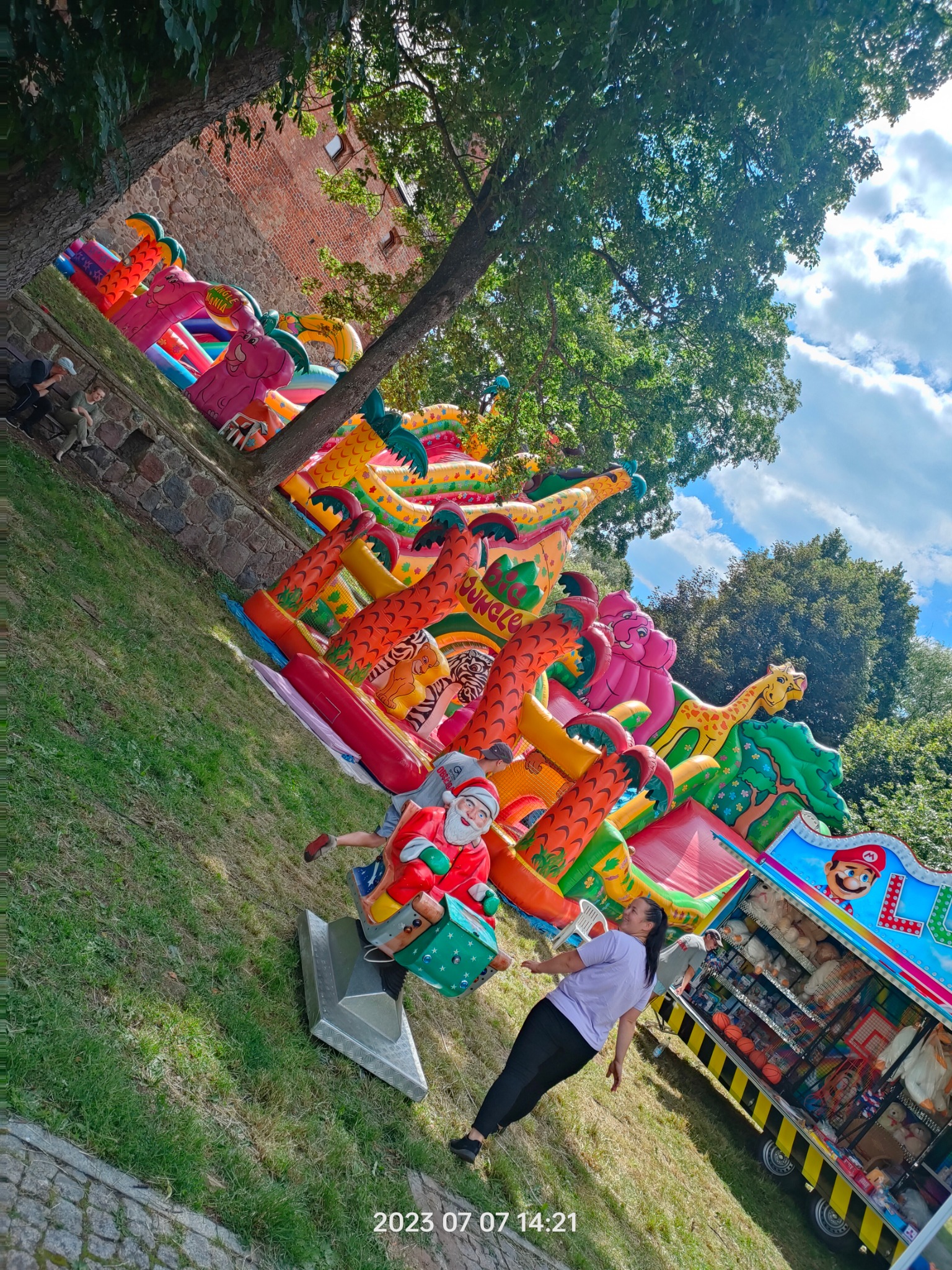 Kolorowy dmuchany plac zabaw w motywie dżungli z postaciami zwierząt, karuzela z motywem świętego mikołaja i budka z zabawkami na trawiastym terenie w słoneczny dzień.