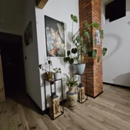 Wnętrze z drewnianą podłogą, ceglanym filarem, obrazem i roślinami w doniczkach na metalowych stojakach oraz ozdobnym wazonem.