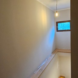 M&M Remonty - Klatka schodowa z drewnianymi schodami i świeżo pomalowanymi ścianami w odcieniach beżu, widoczne listwy przypodłogowe i okno z naturalnym światłem.