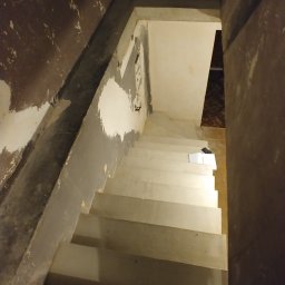 M&M Remonty - Klatka schodowa w trakcie remontu: ściany z resztkami starej farby i gładzi, surowe schody przygotowane do malowania, widoczne ślady po instalacjach elektrycznych.