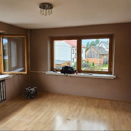 M&M Remonty - Pokój z dwoma oknami o drewnianych ramach, świeżo pomalowanymi ścianami w odcieniach beżu, parkietem na podłodze i starym grzejnikiem pod oknem po lewej stronie. Na parapecie pod oknem po prawej...