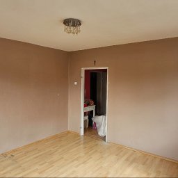 M&M Remonty - Pokój z brązowymi ścianami, drewnianą podłogą i widocznym oknem po prawej stronie. Na suficie wisi kryształowy żyrandol, a w tle widać otwarte drzwi do innego pomieszczenia.