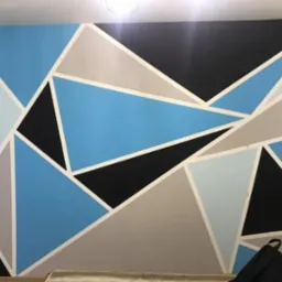Ściana pomalowana w geometryczne wzory z użyciem taśmy malarskiej i farb w odcieniach niebieskiego, szarego i czarnego, widoczny fragment łóżka i plecak w dolnej części kadru.