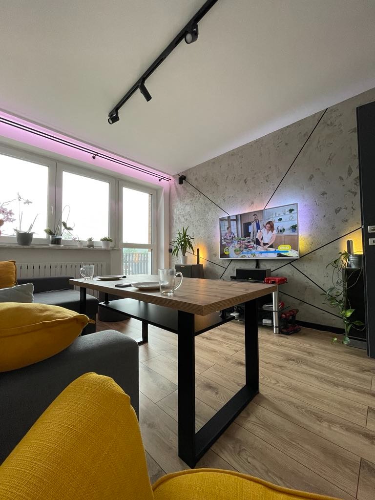 Nowoczesny salon z szarą kanapą, żółtymi poduszkami, drewnianym stolikiem kawowym i ścianą dekoracyjną z telewizorem, oświetlony lampami sufitowymi i listwą LED nad oknem.