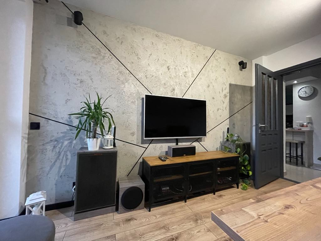 Szara ściana z efektem betonu i geometrycznymi czarnymi liniami, telewizor, drewniana podłoga i meble w salonie.